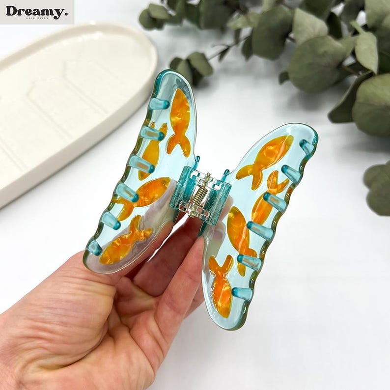 May include: A blue butterfly-shaped hair clip with a gold fish design. The clip has a spring-loaded mechanism to hold hair in place.