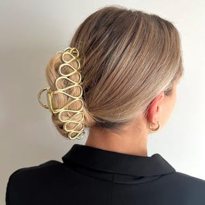 May include: Gold-coloured hair claw clip with an open, abstract design. The clip is holding up blonde hair styled in a bun. The word "Dreamy" is in the top left corner.