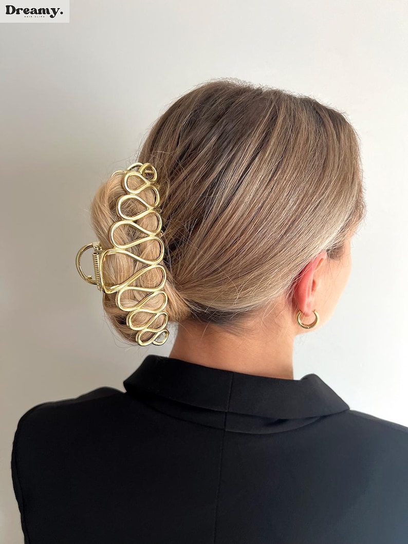 May include: Gold-coloured hair claw clip with an open, abstract design. The clip is holding up blonde hair styled in a bun. The word "Dreamy" is in the top left corner.