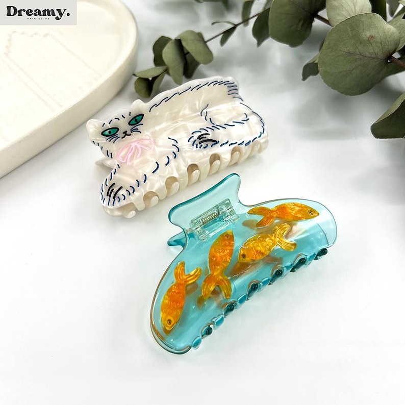 May include: Two hair clips, one with a white cat design and the other with a blue and clear design with gold fish inside.