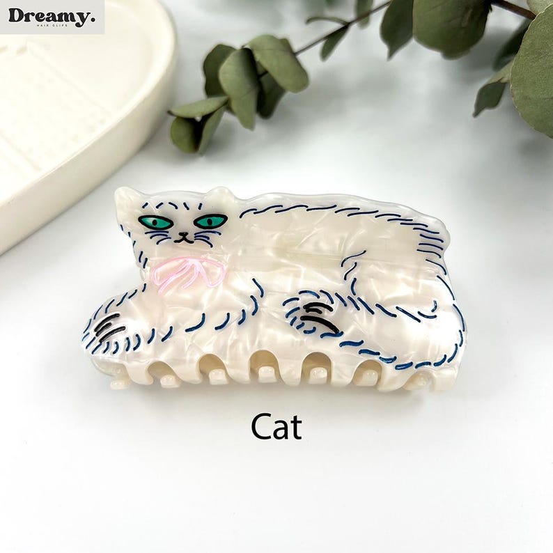 May include: White cat hair clip with blue and black outline. The cat is laying down with a pink bow. The clip is labeled "Cat".