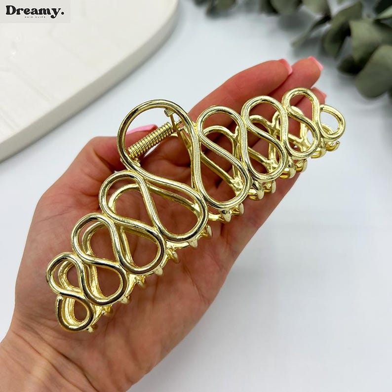 May include: Gold-coloured hair claw clip with an open, looped design. The clip has a spring mechanism and small teeth for grip. The word "Dreamy" is printed in the upper left corner. This hair accessory is designed to hold hair in place.