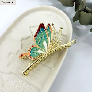 May include: A gold-colored butterfly hair clip with teal, red, and white enamel detailing. The butterfly is perched on a gold-colored branch. The hair accessory is on a white, oval-shaped tray.