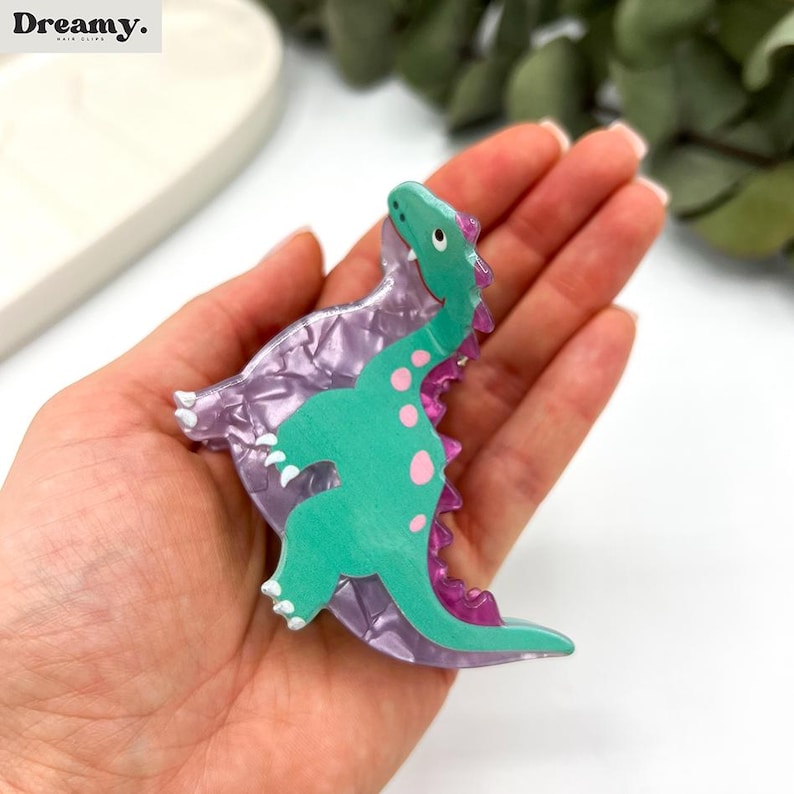 K&ouml;nnte beinhalten: Eine gr&uuml;ne Dinosaurier-Haarklammer mit rosa Punkten und einer lila schimmernden Basis. Die Klammer wird in einer Hand gehalten.