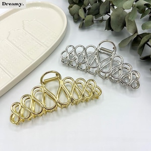 May include: Two metal hair claw clips. One is gold-toned, the other is silver-toned. Both have an open, looped design. The clips are on a white surface, with a white decorative tray and greenery in the background.