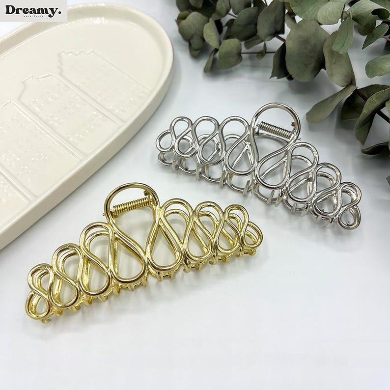 May include: Two metal hair claw clips. One is gold-toned, the other is silver-toned. Both have an open, looped design. The clips are on a white surface, with a white decorative tray and greenery in the background.