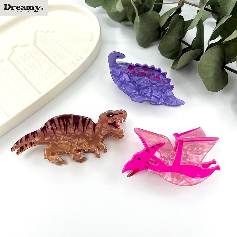 K&ouml;nnte beinhalten: Drei Dinosaurier-Haarklammern aus Kunststoff. Ein lila Stegosaurus, ein brauner T-Rex und ein rosa Pterodactylus.