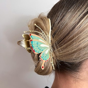 May include: A gold-toned butterfly hair clip with a teal, red, and white wing design. The clip is attached to a bun of blonde hair. The word "Dreamy" is in the top left corner.