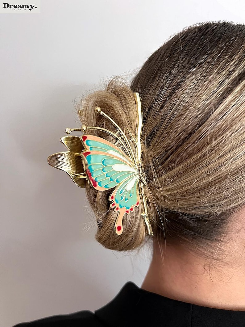 May include: A gold-toned butterfly hair clip with a teal, red, and white wing design. The clip is attached to a bun of blonde hair. The word "Dreamy" is in the top left corner.