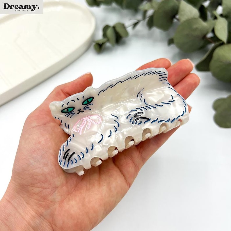 May include: White cat hair clip with blue and green details. The cat is lying down with its paws outstretched.