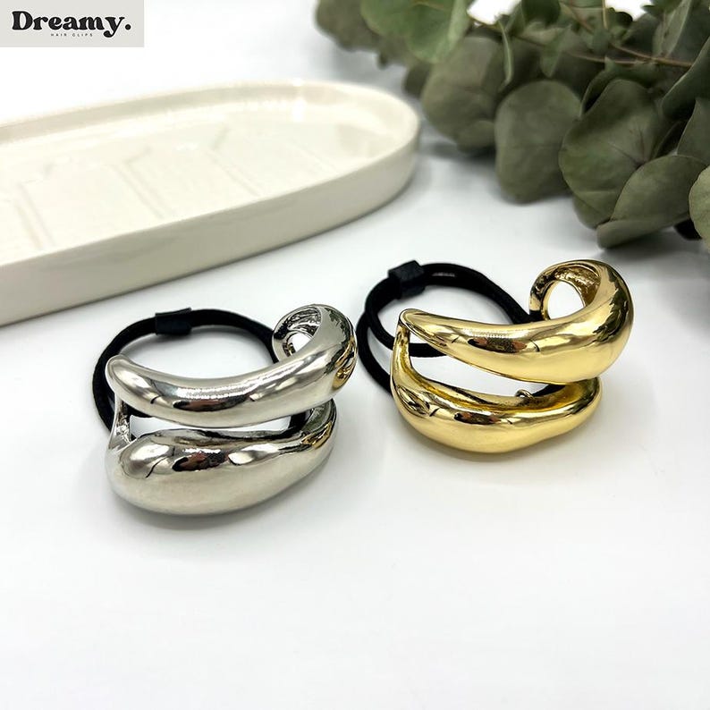 Pu&ograve; includere: Due elastici per capelli con un design astratto dorato e argentato. Gli elastici sono in metallo e hanno un elastico nero.