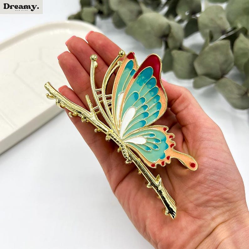 May include: A gold-colored butterfly hair clip with a colorful design. The butterfly wings feature teal, coral, and red accents. The clip is designed to look like the butterfly is perched on a branch.