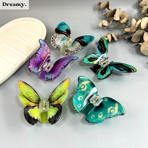 May include: Five butterfly hair clips in shades of purple, green, and teal. Each clip has detailed wing patterns and a clear clasp. The clips are displayed on a light grey surface. The word "Dreamy" is visible in the top left corner.