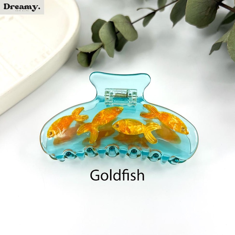 May include: A blue plastic hair claw with a design of four goldfish swimming inside. The claw has a spring-loaded mechanism and a textured grip. The text "Goldfish" is printed below the claw.