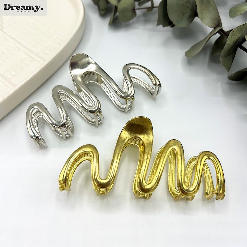 May include: Two metal hair clips in a wavy design. One clip is silver, and the other is gold. The clips are on a white surface with a white tray and greenery in the background. The brand name "Dreamy" is in the top left corner.