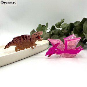 K&ouml;nnte beinhalten: Zwei Plastik-Dinosaurier-Haarklammern. Eine ist ein brauner Tyrannosaurus Rex und die andere ein rosa Pterodactylus.