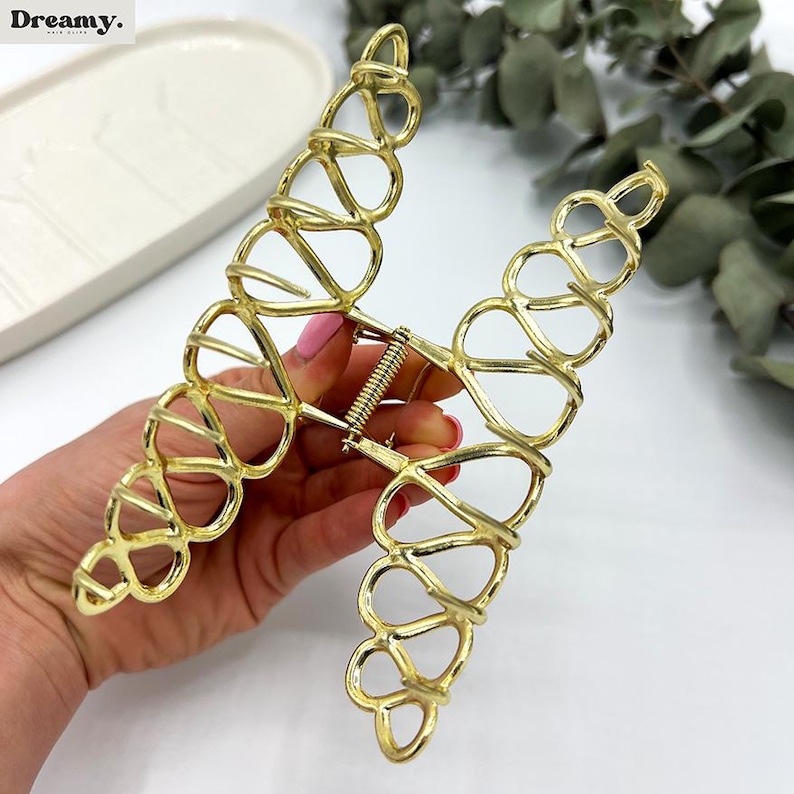 May include: Gold-coloured hair claw clip with an open, heart-shaped design. The clip has a spring mechanism and is made of metal. The clip is designed to hold hair in place and is a stylish accessory.