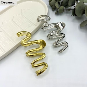May include: Gold and silver-toned metal hair clips in a snake design. The clips are in a wavy, serpentine shape, with a textured finish. The clips are displayed on a white surface with a decorative tray and greenery in the background.
