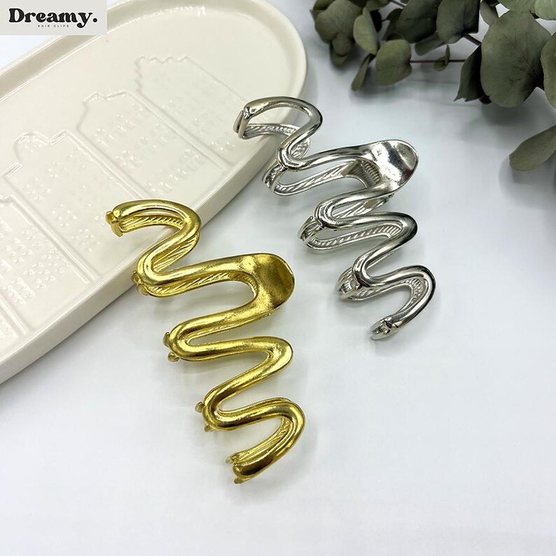 May include: Gold and silver-toned metal hair clips in a snake design. The clips are in a wavy, serpentine shape, with a textured finish. The clips are displayed on a white surface with a decorative tray and greenery in the background.