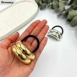 May include: Two gold and silver hair scrunchies with black elastic bands. The scrunchies are made of metal and have a rounded shape.