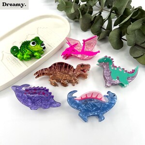 K&ouml;nnte beinhalten: Sechs bunte Dinosaurier-Haarklammern. Die Klammern sind aus Kunststoff und haben eine gl&auml;nzende Oberfl&auml;che. Die Dinosaurier sind ein Cham&auml;leon, ein Pterodactylus, ein T-Rex, ein Stegosaurus, ein Triceratops und ein Velociraptor.