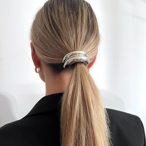 May include: Silver hair clip with a rounded, curved design. The clip is holding a ponytail of blonde hair.