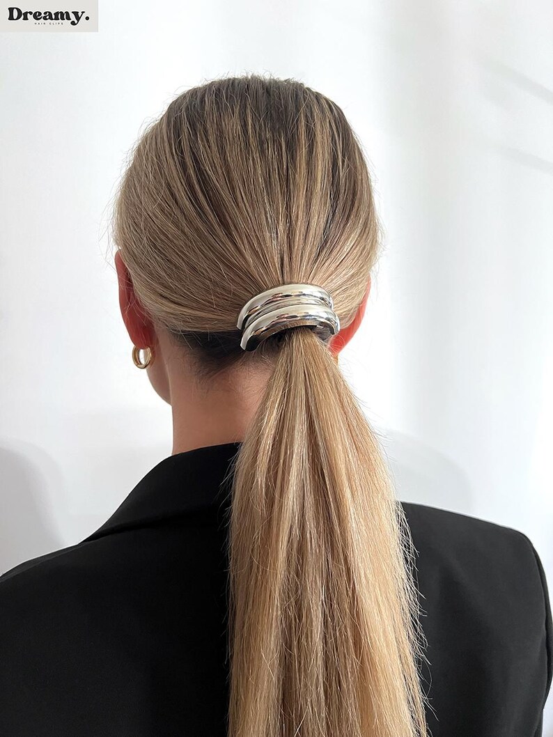 May include: Silver hair clip with a rounded, curved design. The clip is holding a ponytail of blonde hair.