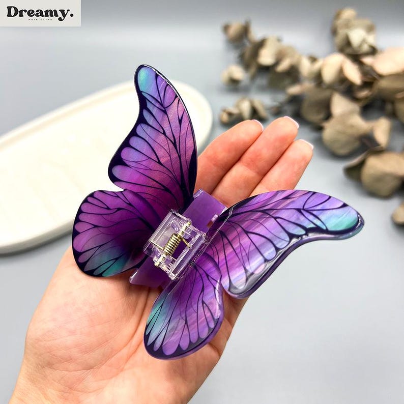 May include: A purple butterfly hair clip with translucent wings. The clip has a clear plastic clasp and a gold-coloured spring. The wings feature a gradient of purple and teal colours, with detailed vein patterns. The brand name "Dreamy" is in the top left corner.