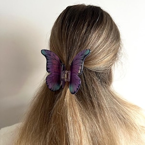 May include: A butterfly hair clip in shades of purple and teal is fastened in blonde hair. The clip is made of translucent plastic and has detailed wing patterns. The word "Dreamy" is printed in the upper left corner.