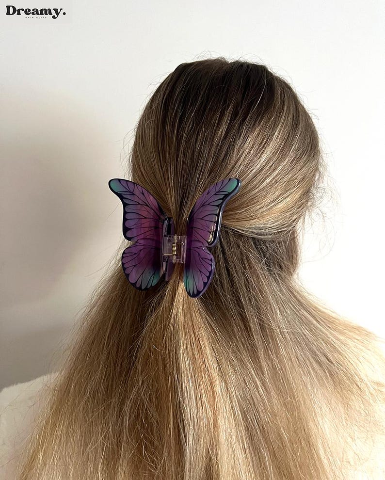 May include: A butterfly hair clip in shades of purple and teal is fastened in blonde hair. The clip is made of translucent plastic and has detailed wing patterns. The word "Dreamy" is printed in the upper left corner.