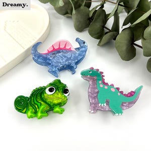 K&ouml;nnte beinhalten: Drei bunte Haarklammern mit einem Cham&auml;leon, einem Stegosaurus und einem Dinosaurier. Die Klammern sind aus Kunststoff und haben einen Marmoreffekt.