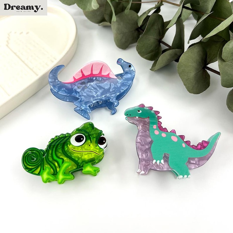 K&ouml;nnte beinhalten: Drei bunte Haarklammern mit einem Cham&auml;leon, einem Stegosaurus und einem Dinosaurier. Die Klammern sind aus Kunststoff und haben einen Marmoreffekt.