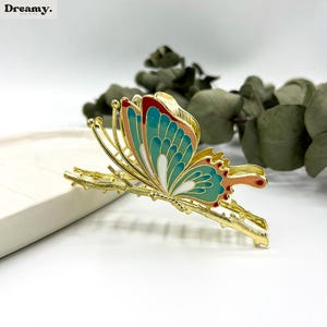 May include: A gold-colored butterfly hair clip with teal, red, and white enamel detailing. The butterfly design is perched on a branch-like structure. The clip is set against a white background with greenery.