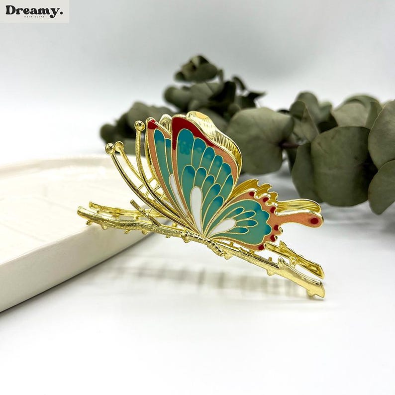 May include: A gold-colored butterfly hair clip with teal, red, and white enamel detailing. The butterfly design is perched on a branch-like structure. The clip is set against a white background with greenery.