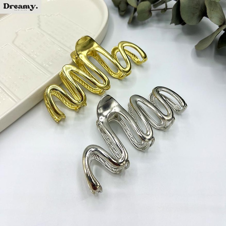 May include: Gold and silver-colored hair clips in a wavy, abstract design. The clips are made of metal and have a shiny finish. The clips are arranged on a white surface, with a white tray in the background.