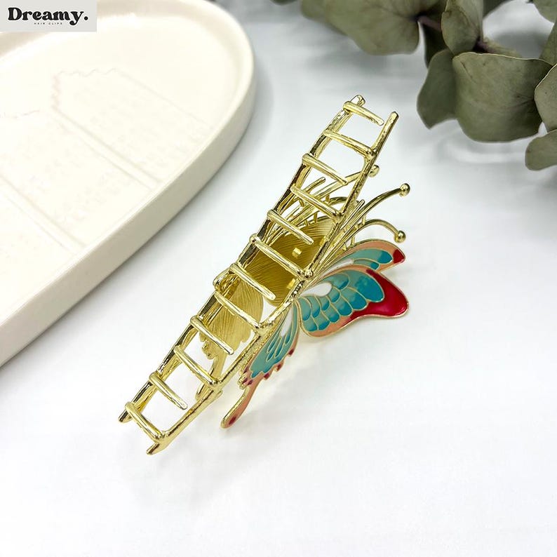 May include: Gold-toned metal hair claw clip with a butterfly design. The butterfly wings are painted with teal, red, and peach enamel. The clip has a rectangular shape with a ladder-like design.