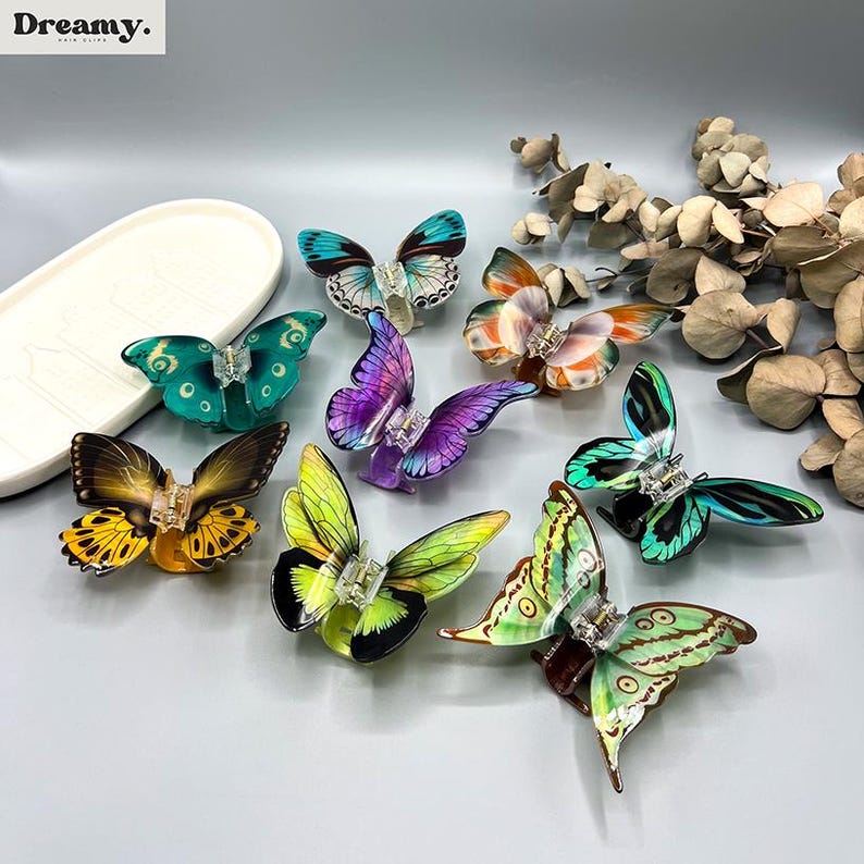 May include: A selection of butterfly hair clips in shades of blue, green, yellow, purple, and orange. Each clip has detailed wing patterns and a clear clasp. The word "Dreamy" is visible in the top left corner.