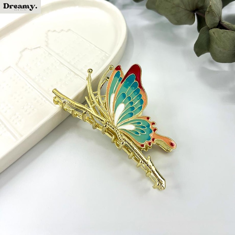 May include: A gold-colored butterfly hair clip with teal, red, and orange accents. The butterfly is perched on a gold-colored branch. The hair clip is on a white surface.
