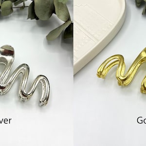 May include: Silver and gold-colored hair clips with a wavy design. The silver clip is on the left, and the gold clip is on the right. The word "Silver" is below the silver clip, and the word "Gold" is below the gold clip.