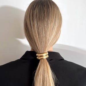 May include: A gold metal hair tie with a double ring design. The hair tie is holding a ponytail of blonde hair.
