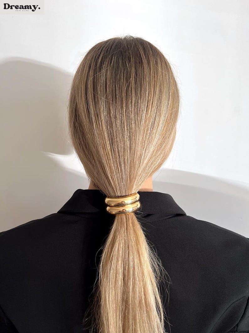 May include: A gold metal hair tie with a double ring design. The hair tie is holding a ponytail of blonde hair.