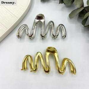 May include: Two metal hair clips in a wavy design. One clip is silver, and the other is gold. The clips have a smooth, shiny finish and are designed to hold hair in place. The clips are on a white surface.