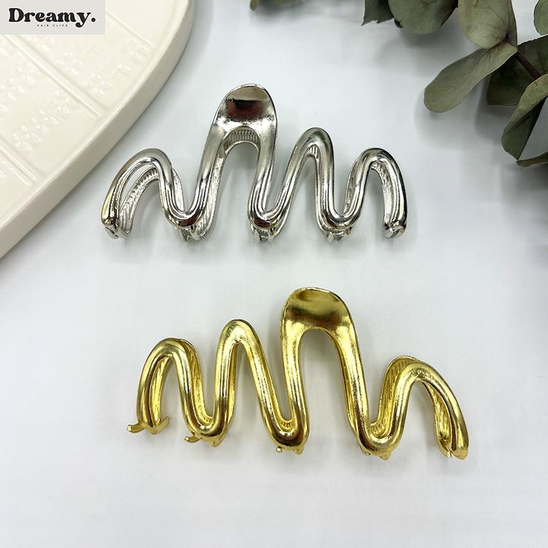 May include: Two metal hair clips in a wavy design. One clip is silver, and the other is gold. The clips have a smooth, shiny finish and are designed to hold hair in place. The clips are on a white surface.