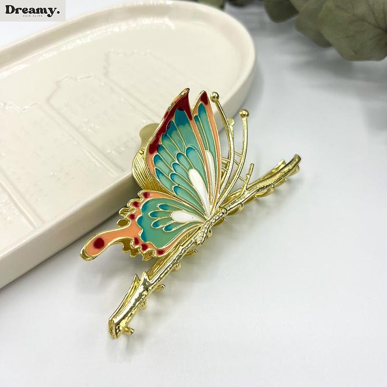 May include: A gold-colored butterfly hair clip with teal, coral, and red enamel wings. The butterfly is perched on a gold branch. The word "Dreamy" is in the top left corner.