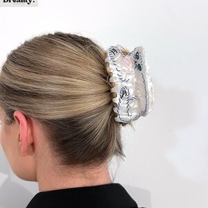 May include: White plastic hair claw with a cat design. The cat is embroidered in blue and pink thread with green eyes.