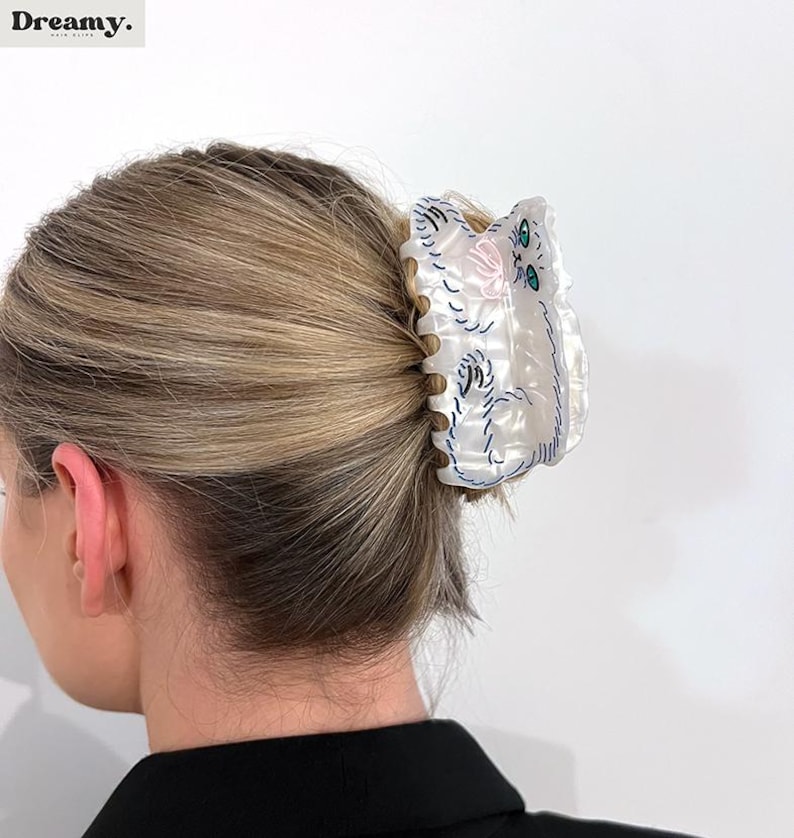 May include: White plastic hair claw with a cat design. The cat is embroidered in blue and pink thread with green eyes.