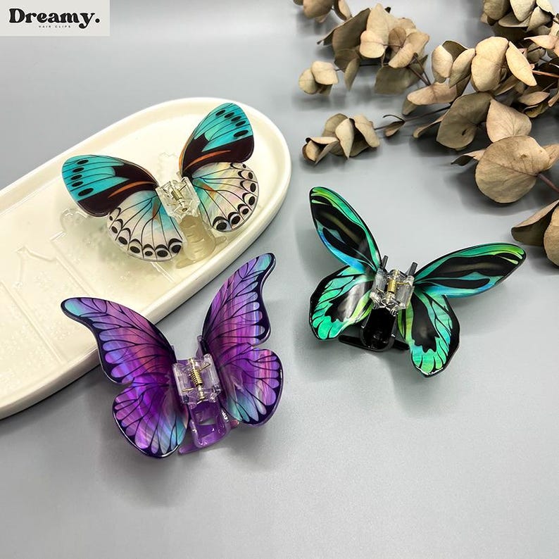 May include: Three butterfly hair clips in teal, purple, and green. Each clip has a clear plastic clasp. The clips are displayed on a neutral surface. The Dreamy logo is in the top left corner.