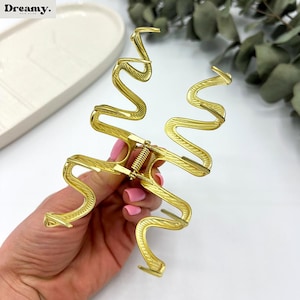 May include: Gold-colored hair claw clip in a unique, wavy design. The clip has a spring mechanism and textured detailing. The Dreamy. logo is visible in the top left corner. A stylish hair accessory.