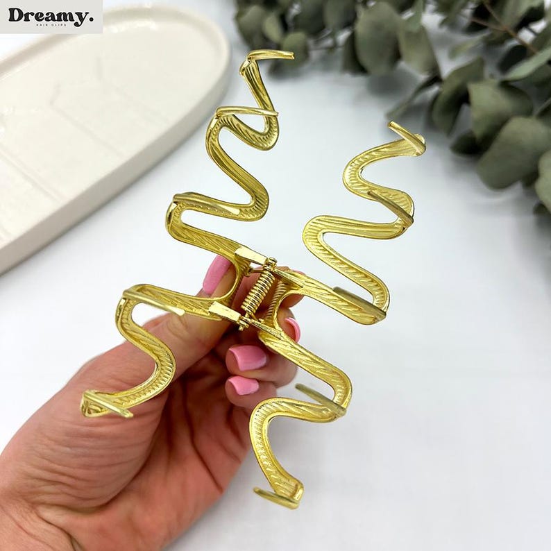 May include: Gold-colored hair claw clip in a unique, wavy design. The clip has a spring mechanism and textured detailing. The Dreamy. logo is visible in the top left corner. A stylish hair accessory.