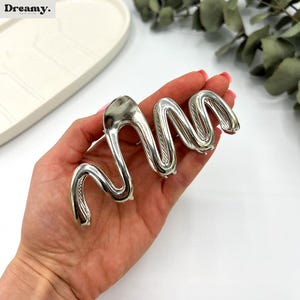 May include: A silver-colored hair clip in a wavy, abstract design. The clip is held in a hand, showcasing its polished, metallic finish. The clip's design is reminiscent of a stylized snake or wave, adding a modern touch to hair accessories.